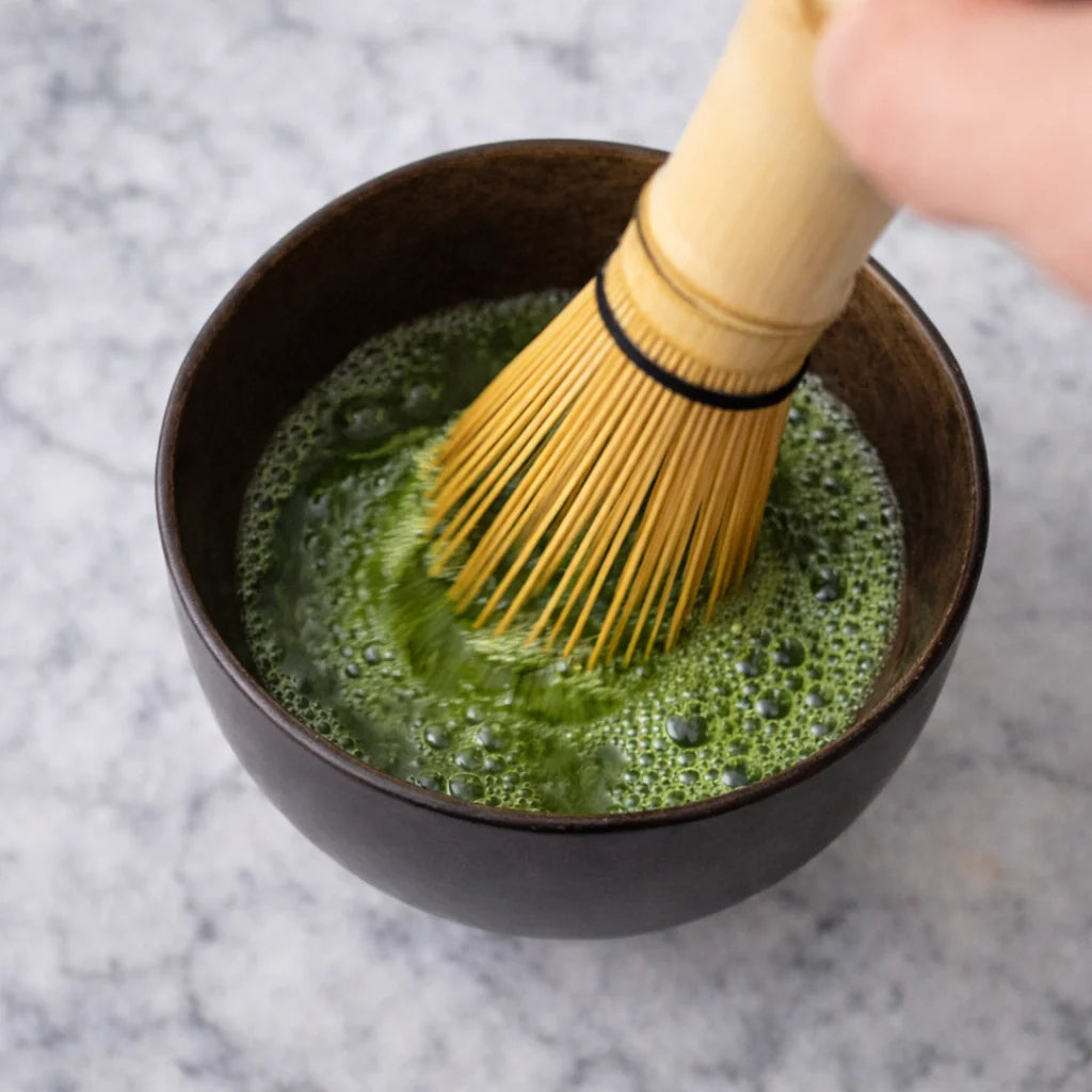 Whisking matcha with a bamboo chasen whisk in a dark ceramic bowl