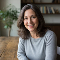 Woman (popcha customer) in a gray sweater sitting at a wooden table with a blurred bookshelf and plant in the background